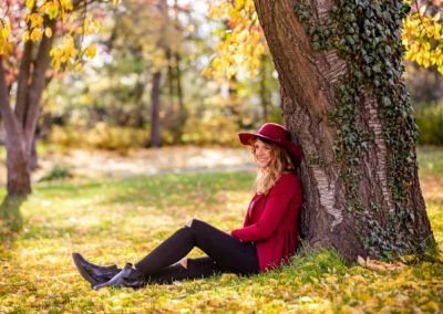 Frau mit rotem Hut sitzt an einen großen Baum gelehnt im Herbstlaub